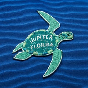 Pu&ograve; includere: Toppa ricamata con un design di tartaruga marina nei toni del verde acqua e verde, con cuciture bianche. Il testo "JUPITER FLORIDA" &egrave; ricamato sul guscio della tartaruga. La toppa &egrave; su uno sfondo blu strutturato.