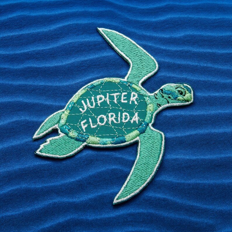 Pu&ograve; includere: Toppa ricamata con un design di tartaruga marina nei toni del verde acqua e verde, con cuciture bianche. Il testo "JUPITER FLORIDA" &egrave; ricamato sul guscio della tartaruga. La toppa &egrave; su uno sfondo blu strutturato.