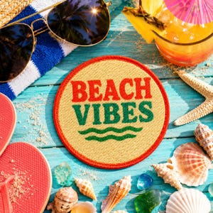May include: A circular embroidered patch with a tan background and a red border. The text BEACH VIBES is stitched in red and green, with two green wavy lines underneath. The patch is surrounded by sunglasses, a drink, and various seashells.