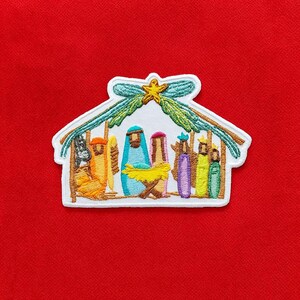 May include: An embroidered patch featuring a nativity scene under a thatched roof with a yellow star at the peak. The design includes figures in blue, orange, purple, yellow, and green robes, with a yellow manger centred on a white background.