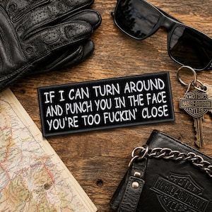 May include: A rectangular black embroidered patch with white text that reads: IF I CAN TURN AROUND AND PUNCH YOU IN THE FACE YOU'RE TOO FUCKIN' CLOSE. The patch is displayed alongside black leather gloves, sunglasses, a map, and a metal keychain.