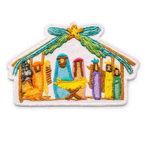 May include: An embroidered patch featuring a nativity scene inside a stable structure. The design includes figures in orange, blue, pink, purple, yellow, and green threads, with a yellow star at the top and green foliage accents on a white background.