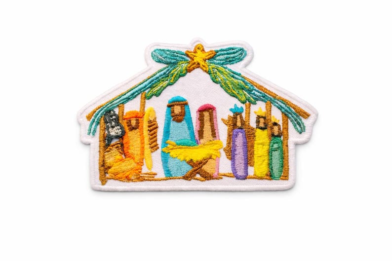 May include: An embroidered patch featuring a nativity scene inside a stable structure. The design includes figures in orange, blue, pink, purple, yellow, and green threads, with a yellow star at the top and green foliage accents on a white background.