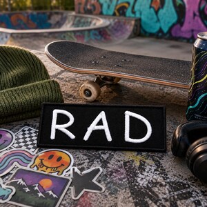 May include: A rectangular black patch with the white embroidered text RAD, positioned near an olive green knit beanie, a skateboard, a black energy drink can with colourful wavy lines, black headphones, and several colourful stickers.