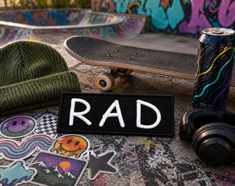 Pode incluir: Um emblema retangular preto com o texto bordado em branco RAD, posicionado perto de um gorro de malha verde-oliva, um skate, uma lata de bebida energética preta com linhas onduladas coloridas, auscultadores pretos e vários autocolantes.