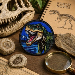 May include: A round, embroidered patch featuring a green and yellow Tyrannosaurus Rex with an open mouth against a blue background. The patch is surrounded by a black border. Other items include a fossilized skull, a notebook with "FIELD NOTES" and a dinosaur sketch, and a magnifying glass.