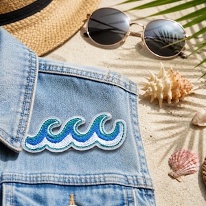 May include: A denim jacket with a blue and white wave patch. The image also includes a straw hat, sunglasses, seashells, and a rope on a sandy surface, evoking a beach theme.