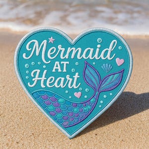 May include: A heart-shaped patch with the words "Mermaid at Heart" in white script. The patch is teal with a mermaid tail design in shades of purple and blue, and small pink hearts. The patch is on a sandy beach.