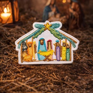 May include: Embroidered patch showing a nativity scene within a white house-shaped frame. The scene includes figures in colourful robes, a star, and a teal and green bow. The patch is set against a blurred background of a lantern and figures.
