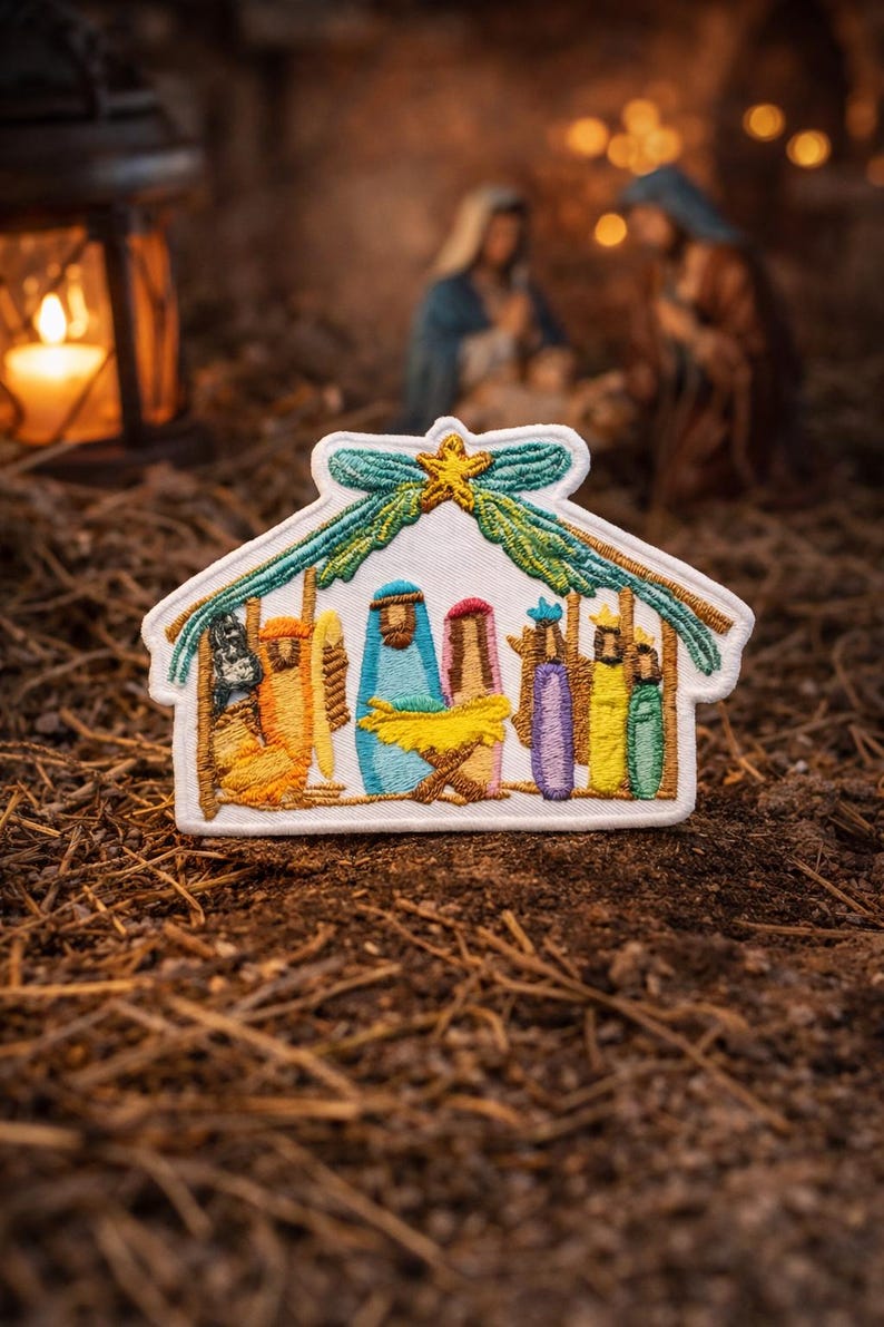May include: A white embroidered patch featuring a nativity scene inside a stable structure. The design includes figures in orange, blue, pink, purple, yellow, and green, with a yellow star at the top centre and green foliage accents above the roof.