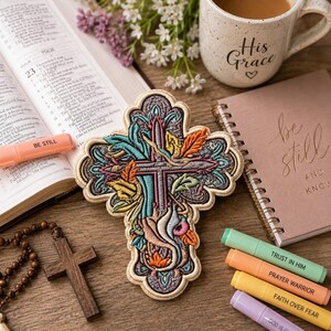 May include: An embroidered cross patch featuring intricate floral and leaf patterns in shades of teal, orange, and purple with a cream-colored border. The design is detailed with multi-colored thread work, creating a textured, decorative religious emblem.