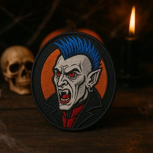 May include: Embroidered patch featuring a vampire with blue hair, red eyes, and fangs. The patch has a black border and an orange background. The vampire is wearing a black jacket and a red shirt. The patch is round and suitable for Halloween.
