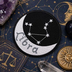 Puede incluir: Parche redondo negro con una luna creciente blanca y estrellas bordadas que forman la constelación de Libra. La palabra "Libra" está bordada en escritura gris. El parche está sobre una superficie azul oscuro con otros elementos astrológicos.