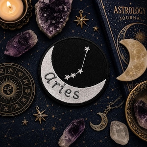 May include: A circular black embroidered patch featuring a silver crescent moon and the word "aries" in lowercase letters. Above the moon, a constellation of five small white stars is stitched onto the black fabric background.