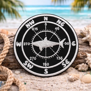 Puede incluir: Parche bordado en blanco y negro con un diseño de brújula y un tiburón en el centro. La rosa de los vientos indica direcciones como NO, NE, SE y SO. El parche es redondo y tiene un tema náutico.