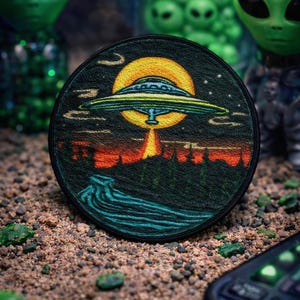 May include: Round embroidered patch featuring a UFO over a landscape with a yellow moon. The patch has a black border and is set against a backdrop of sand and small green rocks. Green alien figures are in the background.