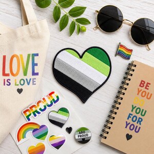 May include: A collection of pride-themed items including a canvas tote bag with the text LOVE IS LOVE, a heart-shaped aromantic flag patch, round sunglasses, a rainbow flag pin, a spiral notebook with the text BE YOU DO YOU FOR YOU, and stickers.