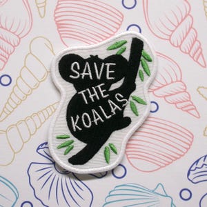 May include: Black and white embroidered patch of a koala holding onto a branch with green leaves. The text "SAVE THE KOALAS" is embroidered in white on the patch.