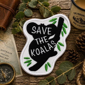 May include: A white embroidered patch with a black silhouette of a koala climbing a tree branch. The patch features green leaves and the white text "SAVE THE KOALAS" stitched across the center of the koala figure, finished with a white border.