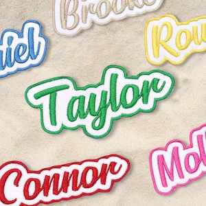 May include: Embroidered name patches in various colours, including green, red, blue, and pink, with white outlines. The names are in a cursive font. The patches are displayed on a sandy surface.