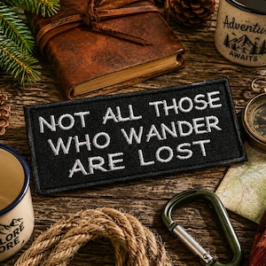 Op de afbeelding: Een rechthoekige zwarte geborduurde patch met de witte tekst "NOT ALL THOSE WHO WANDER ARE LOST" in een strak schreefloos lettertype. De patch heeft een gestikte rand en is ontworpen om te bevestigen op stoffen items zoals tassen of jassen.