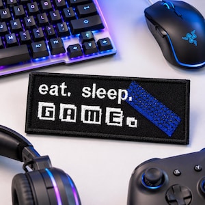 May include: A rectangular black embroidered patch featuring the white text "eat. sleep." above the word "GAME." in a pixelated font. A diagonal blue textured strip is stitched across the upper right corner of the patch.