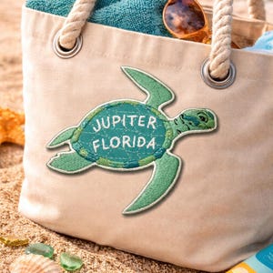 Pu&ograve; includere: Una borsa tote in tela beige con manici in corda, con una toppa a forma di tartaruga verde con la scritta "JUPITER FLORIDA". La borsa contiene un asciugamano turchese e occhiali da sole. La scena &egrave; ambientata su una spiaggia sabbiosa con conchiglie e vetro marino.