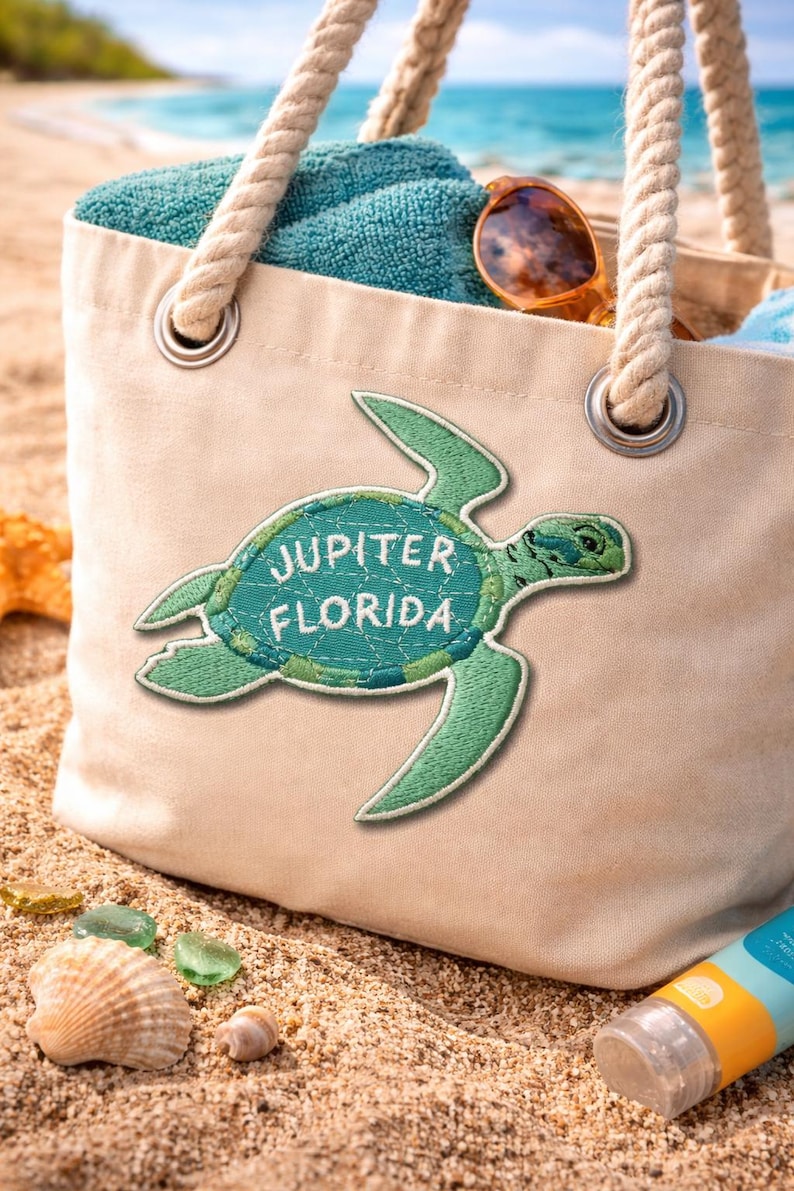 Pu&ograve; includere: Una borsa tote in tela beige con manici in corda, con una toppa a forma di tartaruga verde con la scritta "JUPITER FLORIDA". La borsa contiene un asciugamano turchese e occhiali da sole. La scena &egrave; ambientata su una spiaggia sabbiosa con conchiglie e vetro marino.