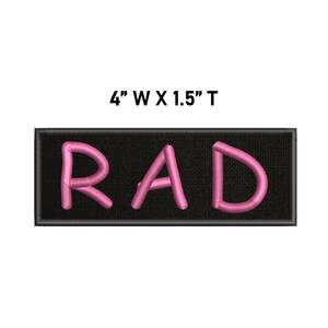 May include: A rectangular black embroidered patch featuring the pink stitched text RAD. The patch has a black border and measures 10.16 cm wide by 3.81 cm tall as indicated by the text above the item.