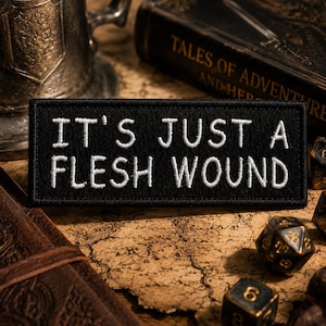 May include: A rectangular black embroidered patch featuring the white stitched text IT'S JUST A FLESH WOUND in a clean sans-serif font. The patch has a finished black border and is designed for attachment to fabric surfaces with its flat rectangular shape.