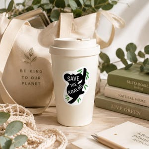 May include: A cream-colored travel mug with a black and white sticker that says "Save the Koalas." A canvas tote bag with the text "Be Kind to Our Planet" is visible in the background. The image also includes books and greenery.
