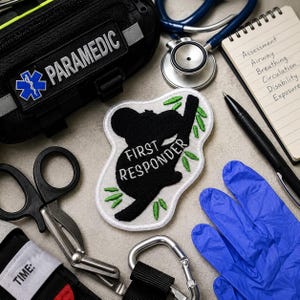 May include: A white embroidered patch featuring a black silhouette of a koala climbing a branch with green leaves and the text FIRST RESPONDER in white. The patch is surrounded by medical gear including a black bag, stethoscope, notepad, and gloves.