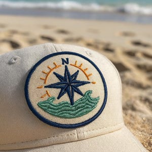 May include: A beige baseball cap with a circular embroidered patch featuring a compass rose design. The patch includes a navy blue star, orange sun rays, and green waves. The background shows a sandy beach and ocean.