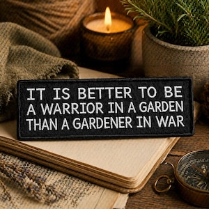 May include: A black rectangular patch with white text: "IT IS BETTER TO BE A WARRIOR IN A GARDEN THAN A GARDENER IN WAR." The patch is on a book, with a lit candle, a small potted plant, and a compass in the background.
