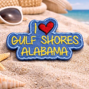 May include: A blue embroidered patch with the text "I LOVE GULF SHORES ALABAMA" in yellow, featuring a red heart. The patch is on a sandy beach with sunglasses, a starfish, and seashells.