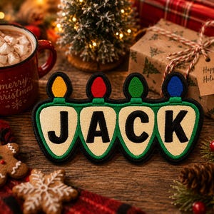 May include: A decorative embroidered patch shaped like four connected Christmas light bulbs, each featuring a letter to spell the name JACK. The bulbs are cream-colored with black letters and topped with yellow, red, green, and blue light caps.
