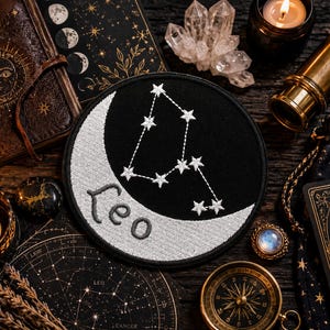 May include: A circular embroidered patch featuring a black background with a white crescent moon and the Leo constellation depicted in white thread. The word Leo is stitched in a cursive font across the lower portion of the crescent moon.