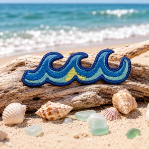 May include: An embroidered wave patch in shades of blue and yellow. The patch, shaped as three waves, rests on driftwood on a sandy beach. Seashells and sea glass are scattered around the driftwood, with the sea in the background.