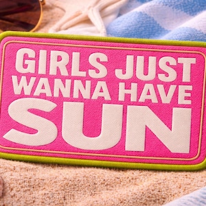 Könnte beinhalten: Ein rechteckiger, pinkfarbener Aufnäher mit dem weißen Text "GIRLS JUST WANNA HAVE SUN". Der Aufnäher hat einen grünen Rand und ist von Muscheln, Sonnenbrillen und einem blau-weiß gestreiften Handtuch auf Sand umgeben.