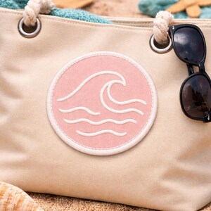 Peut inclure: Sac fourre-tout en toile beige avec un &eacute;cusson circulaire rose repr&eacute;sentant un motif de vagues blanches. Le sac a des poign&eacute;es en corde et des &oelig;illets m&eacute;talliques. Des lunettes de soleil sont pos&eacute;es sur le sac. Th&egrave;me plage.