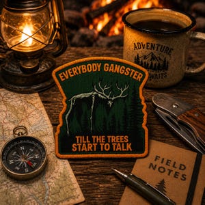 May include: A shield-shaped embroidered patch featuring a dark green forest background with a white skeletal deer silhouette. The text reads EVERYBODY GANGSTER at the top and TILL THE TREES START TO TALK at the bottom in bold orange lettering.