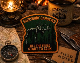 May include: A shield-shaped embroidered patch featuring a dark green forest background with a white skeletal deer silhouette. The text reads EVERYBODY GANGSTER at the top and TILL THE TREES START TO TALK at the bottom in bold orange lettering.