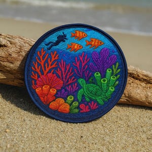 Peut inclure: Écusson rond brodé représentant une scène sous-marine. Le motif comprend un plongeur, des poissons orange, une tortue verte et des coraux colorés. L'écusson a une bordure bleu foncé et est posé sur un fond de plage de sable.