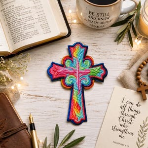 May include: A vibrant, multi-colored embroidered cross patch featuring swirling patterns in shades of pink, green, yellow, and blue, outlined with a bold dark blue border. The cross is positioned centrally, showcasing intricate thread work and texture.
