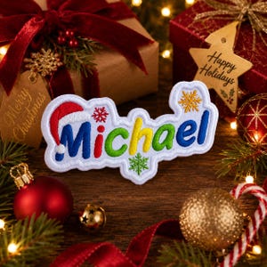 May include: A white embroidered patch featuring the name Michael in multi-colored letters. The letter M is topped with a red and white Santa hat. The design includes small red, green, and gold snowflake accents on a white background.
