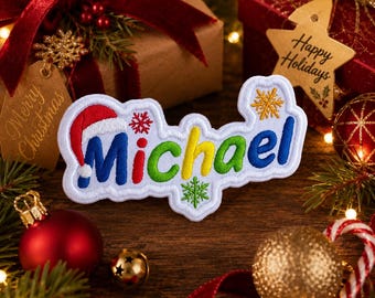 Könnte beinhalten: Ein weißer gestickter Aufnäher mit dem Namen Michael in bunten Buchstaben. Der Buchstabe M ist mit einer rot-weißen Weihnachtsmütze versehen. Das Design enthält kleine rote, grüne und goldene Schneeflocken auf weißem Hintergrund.