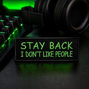 May include: Black gaming setup featuring a keyboard, mouse, and headset. A patch with the text "STAY BACK I DON'T LIKE PEOPLE" is displayed. The keyboard and mouse have green LED lighting.
