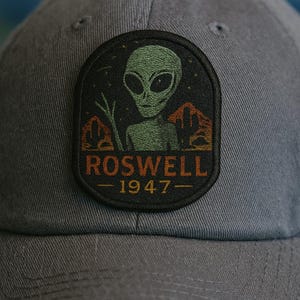 May include: Grey baseball cap with a patch depicting a green alien, desert scenery, and the text "ROSWELL 1947". The patch has black, orange, and green detailing. The cap is made of a textured material.