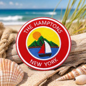 Puede incluir: Parche bordado con borde rojo y las palabras "THE HAMPTONS NEW YORK" en blanco. El centro presenta un círculo amarillo con una escena de montaña, un velero y un sol. El parche está sobre una playa de arena con conchas y madera flotante.
