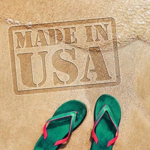 K&ouml;nnte beinhalten: Gr&uuml;ne Flip-Flops mit rosa Riemen liegen auf Sand neben einem Abdruck mit der Aufschrift "MADE IN USA". Das Bild f&auml;ngt ein Sommerthema ein und deutet auf Freizeit und in den USA hergestellte Produkte hin.